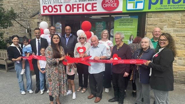 A ribbon-cutting ceremony at New Mill Post Office with a diverse group of people holding balloons and smiling.