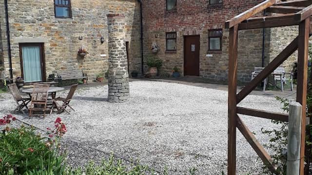 Holiday cottages with stone walls, gravel courtyard, wooden seating, and lush greenery under a blue sky in Yorkshire.