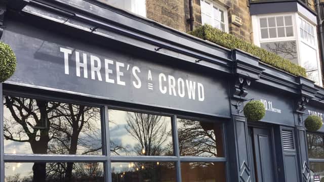 Black exterior of "Three's a Crowd" bar with large windows, greenery accents, and a historic building backdrop.