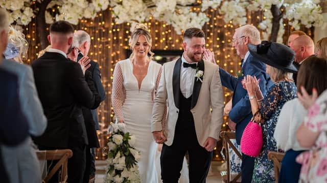 Bride and groom walk hand in hand down an aisle adorned with flowers and fairy lights, surrounded by applauding guests.