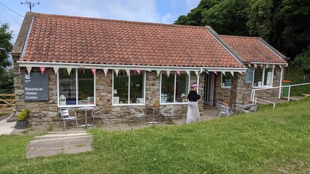Coastal visitor center with stone walls, a red-tiled roof, outdoor seating, and bunting in a green landscape.