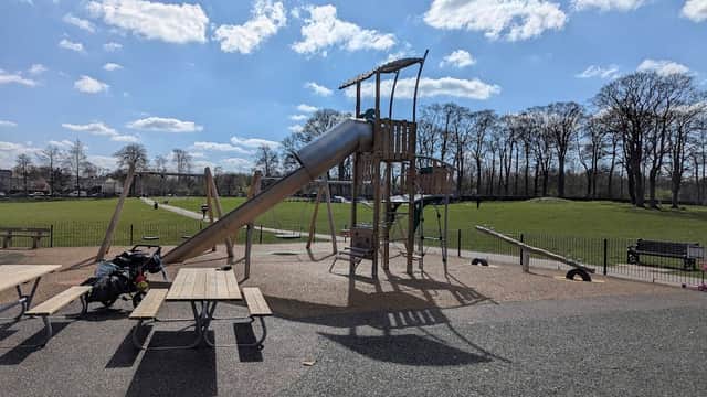 Playground with a slide, picnic tables, and green hills under a blue sky with fluffy clouds in Menston Park.