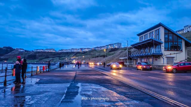 Pay & Display parking near the sea, with cars, pedestrians, and a building under a cloudy blue sky in Scarborough.