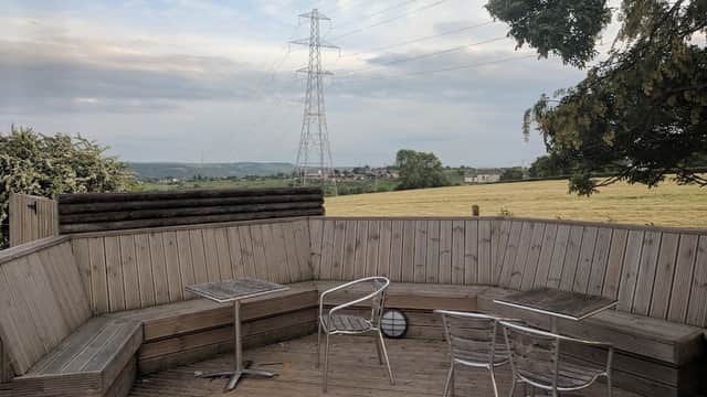 Outdoor seating area with wooden benches and tables, overlooking a grassy field and power lines in the distance.