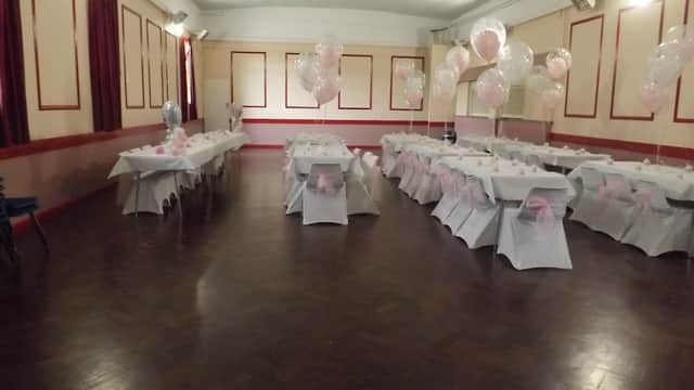 Event venue with tables dressed in white tablecloths and pink bows, decorated with balloons and a polished wooden floor.