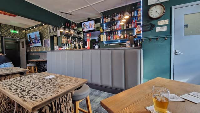 Bar interior with a wooden counter, shelves of drinks, two TVs, and tables with patterned surfaces. A pint of beer sits on a table.
