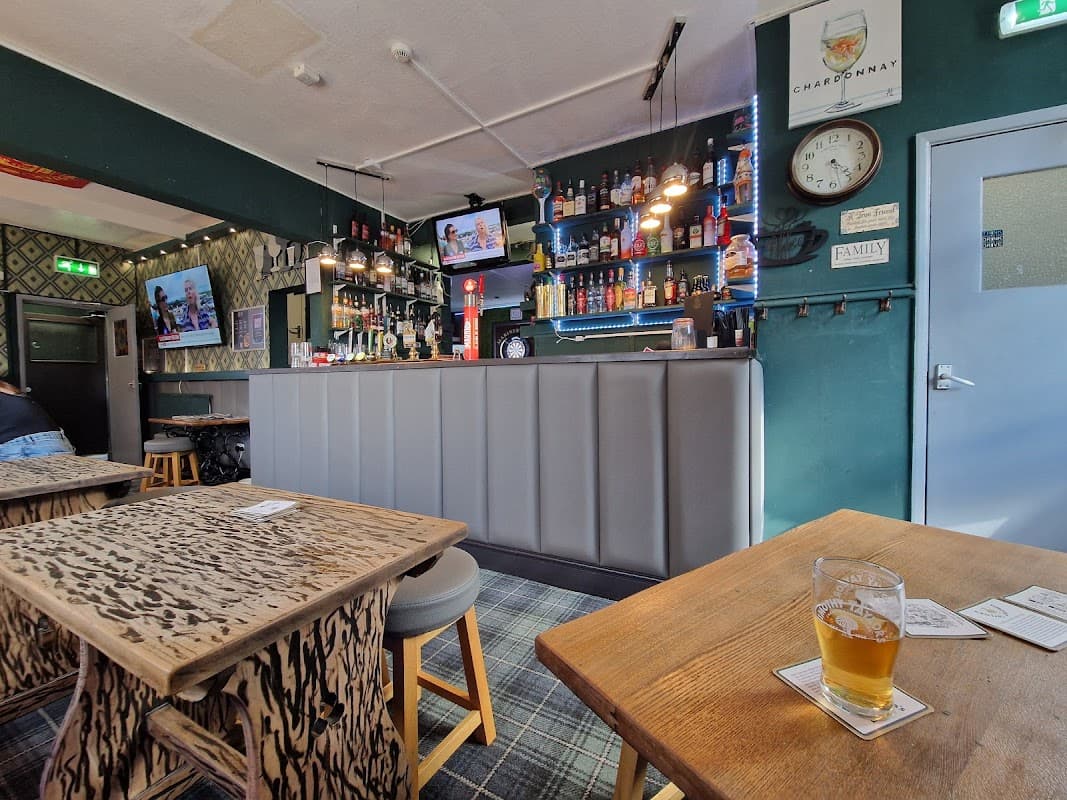 Bar interior with a wooden counter, shelves of drinks, two TVs, and tables with patterned surfaces. A pint of beer sits on a table.