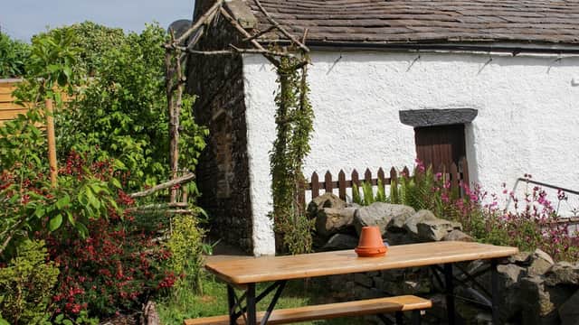 Charming white cottage with a stone wall, wooden door, and rustic garden tables surrounded by colorful flowers and greenery.