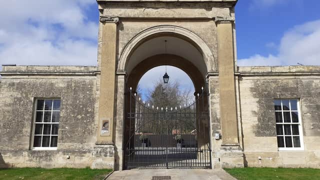 Historic stone archway with wrought iron gates, flanked by large windows and a grassy area under a blue sky.