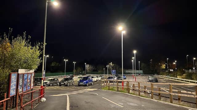 Well-lit car park at night with several parked cars, trees, and a red fence in the foreground.