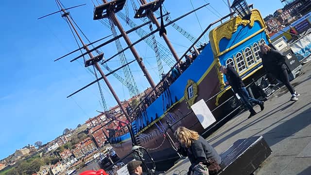 Colorful pirate ship docked at Whitby, with people walking along the waterfront and scenic hills in the background.
