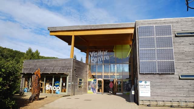 Modern wooden visitor center with large glass windows, solar panels, and people entering under a clear blue sky.
