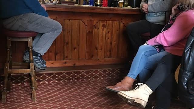 Cozy bar interior with patrons chatting, wooden bar, various drinks displayed, and patterned carpet.