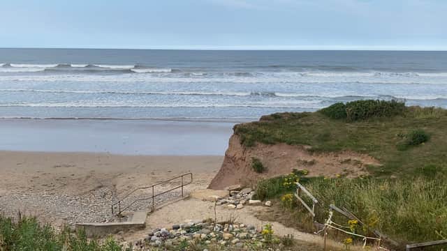 A sandy beach with gentle waves, grassy cliffs, and a rocky path leading to the shoreline under a cloudy sky.