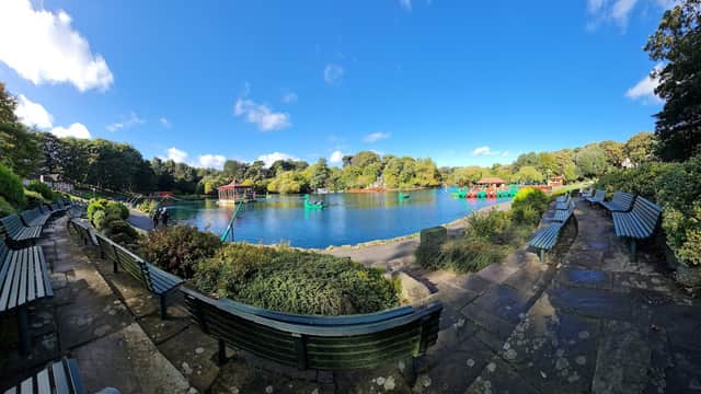Lush greenery surrounds a tranquil lake with colorful boats, benches lining the path under a bright blue sky.