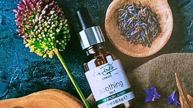 A soothing serum bottle surrounded by flowers, herbs, and wooden bowls on a dark textured surface.