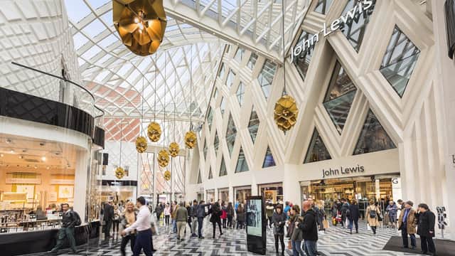 Modern shopping center with a glass roof, geometric patterns on the floor, and shoppers exploring various stores.