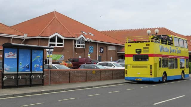 Bright yellow double-decker bus parked outside the Whitby Co-op, with a bus stop and cars in the background.