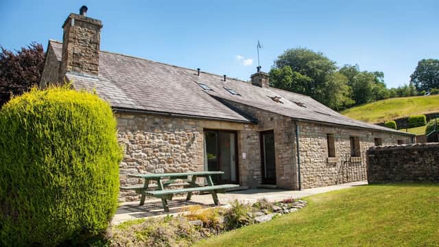 Stone building with a slate roof, picnic table, and lush green lawn surrounded by trees in a sunny landscape.