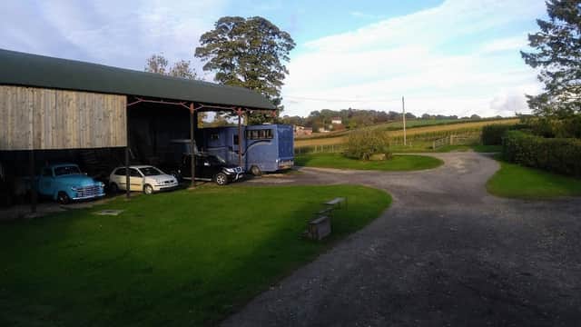 Charming farm holiday cottage with a blue truck, vintage cars, and lush green fields under a clear sky.