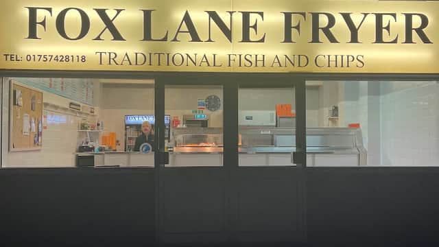 Brightly lit restaurant sign for Fox Lane Fryer, featuring traditional fish and chips, with a staff member visible inside.