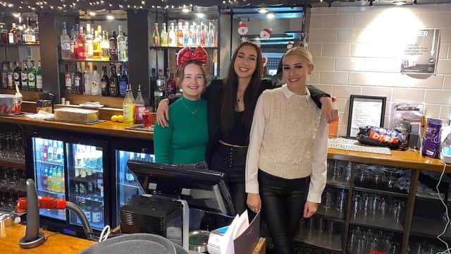 Three women stand behind a bar with shelves of liquor and festive decorations, smiling at the camera.