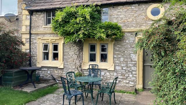 Charming stone cottage with yellow accents, a vine-covered entrance, and a small patio set in a grassy area.