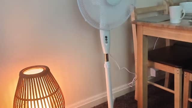 A standing fan next to a woven lamp, with a small table and chair in a cozy room setting.