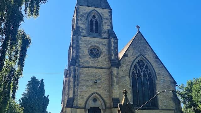 Victorian church with a tall spire, surrounded by gravestones and lush greenery under a clear blue sky.