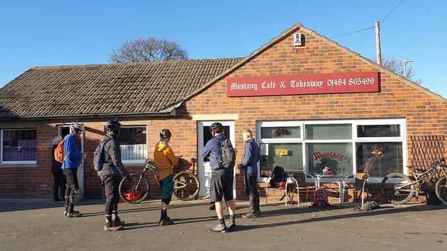 Customers queue outside Mustang Cafe & Takeaway, a brick building with a sign, bicycles parked nearby, on a sunny day.