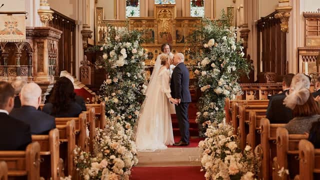 A couple exchanges vows in a decorated church, surrounded by floral arrangements and seated guests.