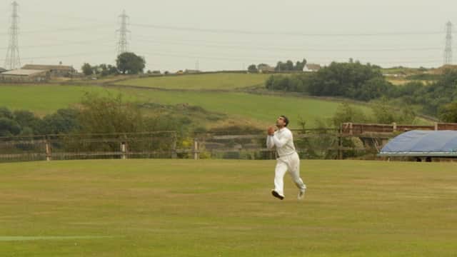 A cricketer in white gear catches a ball on a grassy field with hills and power lines in the background.