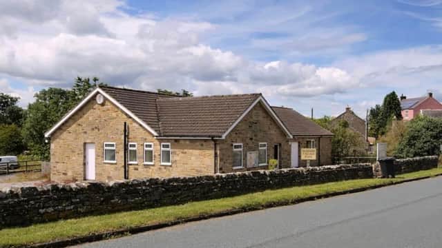 Stone building with a sloped roof, surrounded by greenery and a stone wall, along a quiet road in Harmby, Yorkshire.