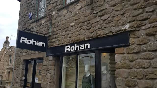 Stone building with large windows, featuring mannequins in outdoor clothing and a sign displaying "Rohan" and a discount offer.