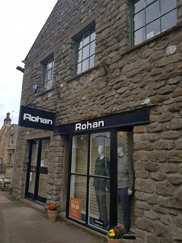Stone building with large windows, featuring mannequins in outdoor clothing and a sign displaying "Rohan" and a discount offer.