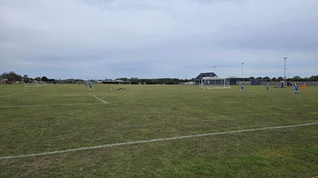 Expansive grassy field with soccer goals, players in blue jerseys, and a pavilion under a cloudy sky.