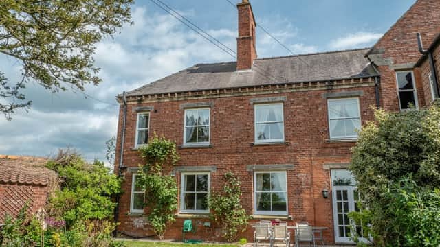 Red brick house with large windows, green garden, patio furniture, and surrounding trees under a partly cloudy sky.