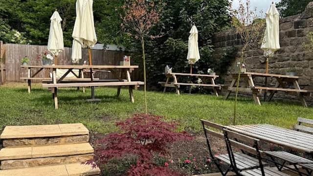 Three picnic tables with umbrellas, surrounded by greenery and a stone wall, with a potted plant in the foreground.