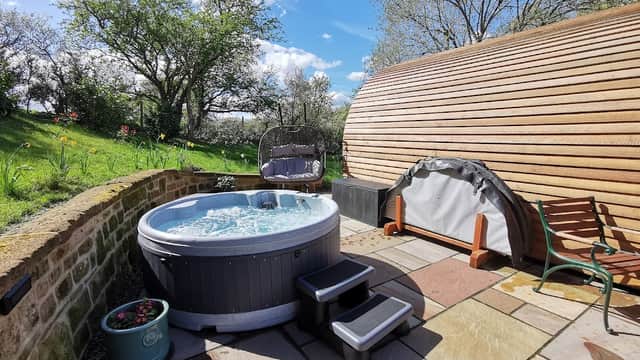 Hot tub with steps surrounded by greenery and trees, alongside a wooden pod structure in a sunny outdoor setting.