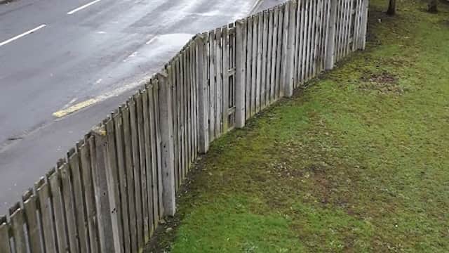 A quiet street with a wooden fence, grass, and a view of distant buildings under a cloudy sky.