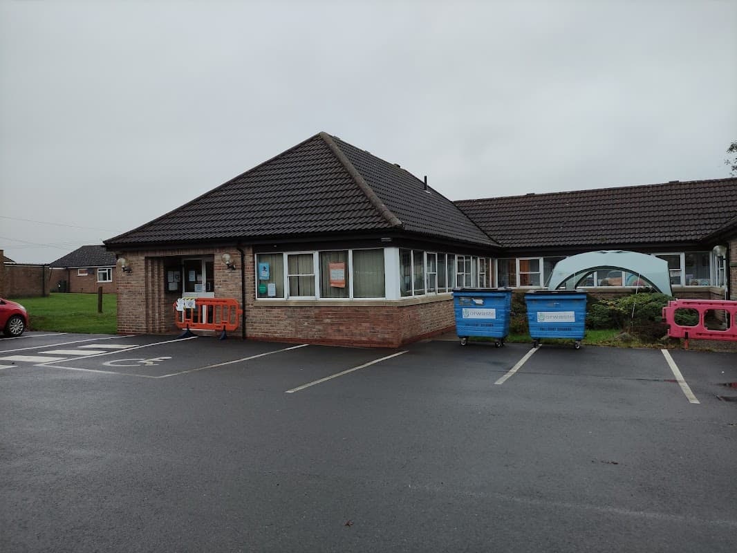 Stillington Surgery building with a sloped roof, parking area, and recycling bins outside on a cloudy day.