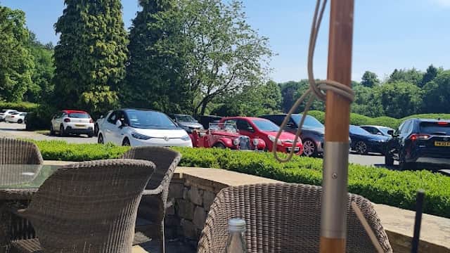 Outdoor seating at a restaurant with a view of parked cars, lush trees, and a sunny blue sky. A drink sits on the table.