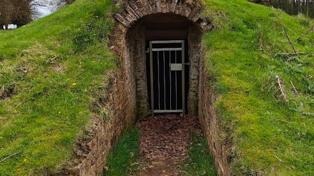 Historic ice house entrance with a grassy mound and a metal gate, surrounded by trees and a dirt path.