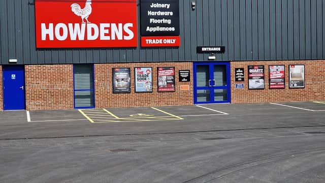 Howdens storefront featuring a red sign, blue entrance doors, and promotional posters on a brick wall.