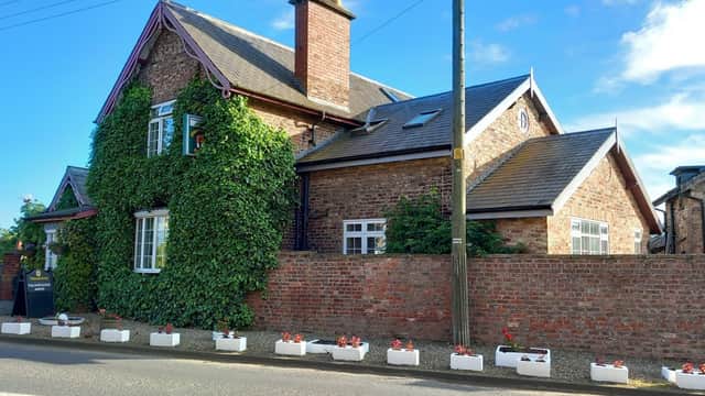Brick building with ivy-covered walls, white window frames, and a flower bed along the front, set against a blue sky.