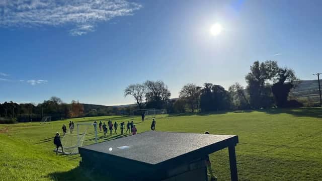 Youth football players practice on a green field under a clear blue sky with the sun shining brightly.