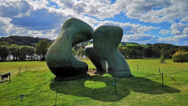 Two large, abstract sculptures resembling figures embrace on a grassy field under a blue sky with fluffy clouds.