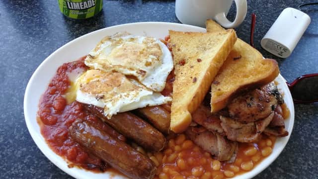 A hearty breakfast plate with sausages, eggs, baked beans, toast, and bacon, alongside a drink and keys.