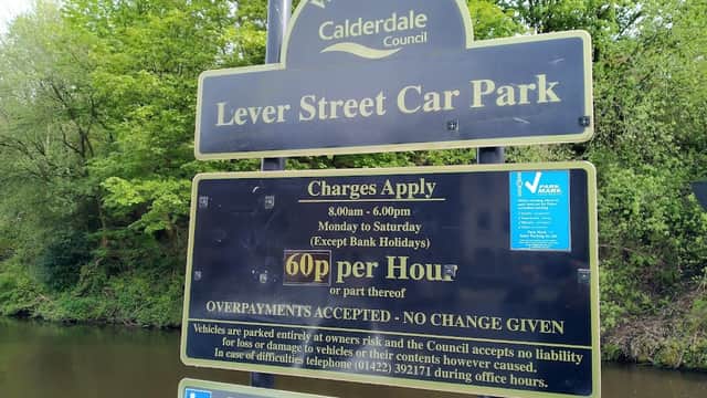Welcome sign for Lever Street Car Park, listing charges and operating hours, surrounded by greenery and a river.