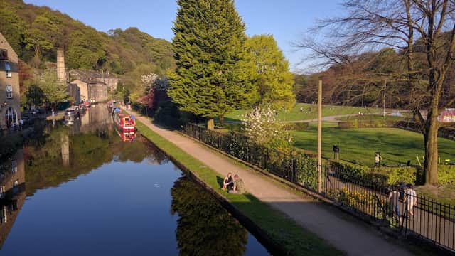 Scenic view of a canal reflecting trees, with people walking along the path and a park in the background in Hebden Bridge.